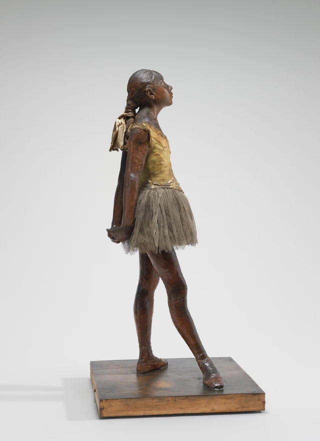 Full-length side view of the same wax sculpture. The forward curve of the chest and abdomen, the lifted chin, and the backward reach of the arms are visible together; her hands clasp low behind the skirt.